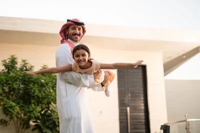 قضاء أوقات ممتعه مع الأهل الأبناء، رجل عربي خليجي سعودي يرتدي الثوب التقليدي والشماغ يرفع ابنته عاليا بإيماءات السعادة، المحافظة على الترابط الأسري، الاعتناء والاهتمام بالاولاد، اللعب والتسلية والترفيه مع العائلة في الهواء الطلق