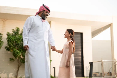 Playing, having fun and entertaining with the family outdoors, spending quality time with parents and children, a Saudi Arabian Gulf man wearing traditional thobe and shemagh playing in the garden with his daughter with happy gestures, maintaining family ties, taking care of and looking after the children