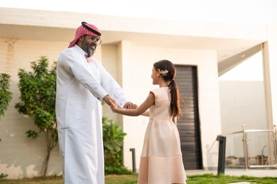 Playing, having fun and entertaining with the family outdoors, spending quality time with parents and children, a Saudi Arabian Gulf man wearing traditional thobe and shemagh playing in the garden with his daughter with happy gestures, maintaining family ties, taking care of and looking after the children