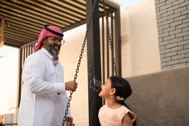 Swinging on a swing, enjoying playing on a sunny day, having a good time outside, a smiling Saudi Arabian Gulf girl playing in the park with expressions of pleasure and happiness with her father
