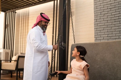 Enjoying playing on a sunny day, having a good time outside, a smiling Saudi Arabian Gulf girl playing in the park with expressions of pleasure and happiness with her father, swinging on the swing