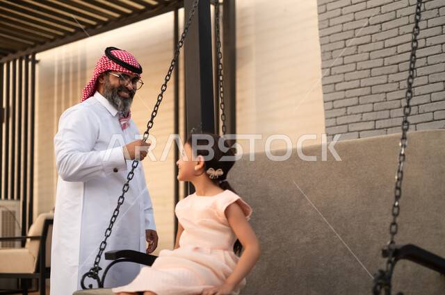 Swinging on a swing, enjoying playing on a sunny day, having a good time outside, a smiling Saudi Arabian Gulf girl playing in the park with expressions of pleasure and happiness with her father
