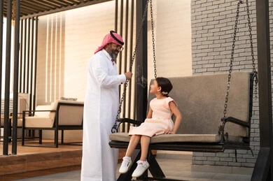 Enjoying playing on a sunny day, having a good time outside, a smiling Saudi Arabian Gulf girl playing in the park with expressions of pleasure and happiness with her father, swinging on the swing
