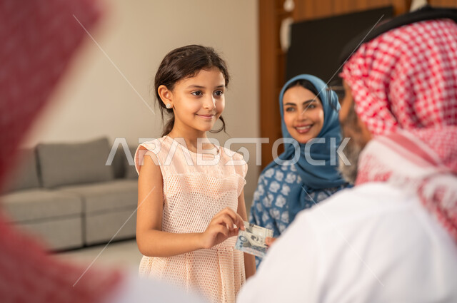 تقديم العيدية في العيد، مفهوم الحب والحنان، دور الأب في حياه الأبناء، جد عربي خليجي سعودي يرتدي الثوب التقليدي والشماغ يجلس على كرسي يعطي الفلوس لحفيدته، عادات وتقاليد الاعياد والمناسبات الدينية، ايماءات السعادة والسرور،  اجواء اسرية مليئة بالفرح