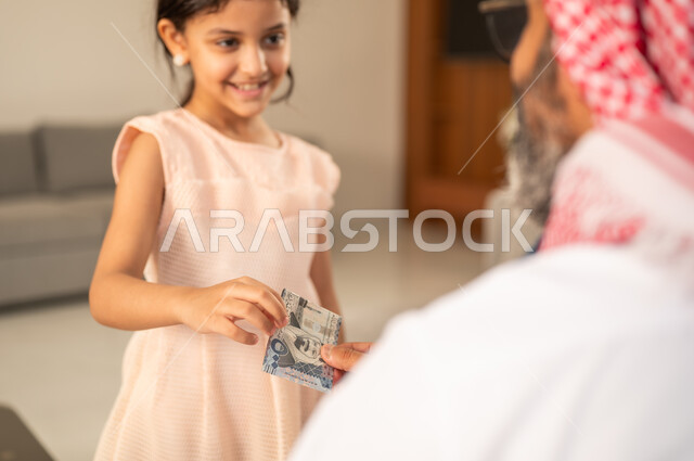 تقديم العيدية في العيد، مفهوم الحب والحنان، دور الأب في حياه الأبناء، جد عربي خليجي سعودي يرتدي الثوب التقليدي والشماغ يجلس على كرسي يعطي الفلوس لحفيدته، عادات وتقاليد الاعياد والمناسبات الدينية، ايماءات السعادة والسرور،  اجواء اسرية مليئة بالفرح