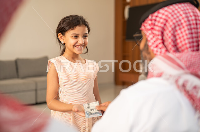 تقديم العيدية في العيد، مفهوم الحب والحنان، دور الأب في حياه الأبناء، جد عربي خليجي سعودي يرتدي الثوب التقليدي والشماغ يجلس على كرسي يعطي الفلوس لحفيدته، عادات وتقاليد الاعياد والمناسبات الدينية، ايماءات السعادة والسرور،  اجواء اسرية مليئة بالفرح