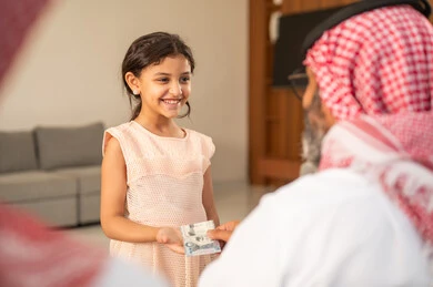 تقديم العيدية في العيد، مفهوم الحب والحنان، دور الأب في حياه الأبناء، جد عربي خليجي سعودي يرتدي الثوب التقليدي والشماغ يجلس على كرسي يعطي الفلوس لحفيدته، عادات وتقاليد الاعياد والمناسبات الدينية، ايماءات السعادة والسرور،  اجواء اسرية مليئة بالفرح