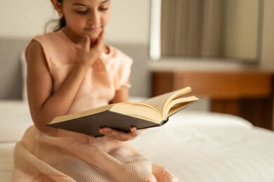 Spending free time learning and self-education, gestures of attention and focus in reading, a Saudi Arabian Gulf girl sitting on the bed reading carefully, reading entertaining picture stories, developing children's reading skills, interest in browsing useful books