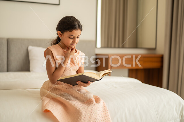 تمضية أوقات الفراغ بالتعلم والتثقف الذاتي, إيماءات الانتباه والتركيز بالمطالعة, فتاة عربية خليجية سعودية تجلس على السرير تقوم بالقراءة بتمعن, مطالعة القصص التصويرية المسلية, تنمية مهارات القراءة للأطفال, الاهتمام بتصفح الكتب المفيدة