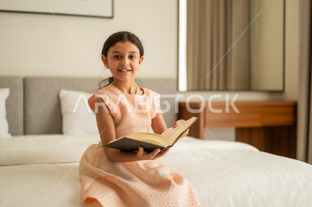 تمضية أوقات الفراغ بالتعلم والتثقف الذاتي, إيماءات الانتباه والتركيز بالمطالعة, فتاة عربية خليجية سعودية تجلس على السرير تقوم بالقراءة بتمعن, مطالعة القصص التصويرية المسلية, تنمية مهارات القراءة للأطفال, الاهتمام بتصفح الكتب المفيدة