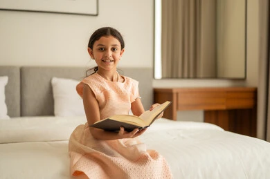 Spending free time learning and self-education, gestures of attention and focus in reading, a Saudi Arabian Gulf girl sitting on the bed reading carefully, reading entertaining picture stories, developing children's reading skills, interest in browsing useful books