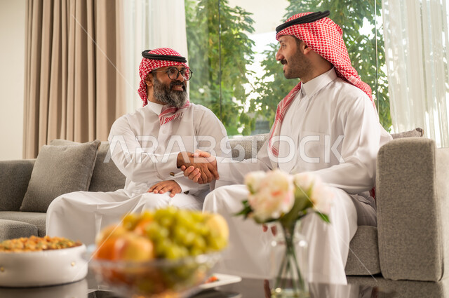 التسامر وتبادل الاحاديث، جلسة عائلية سعيدة، تعابير المودة والمحبة، الاستمتاع بقضاء وقت مع الابناء، رجل كبير بالسن عربي خليجي سعودي يرتدي الثوب التقليدي والشماغ يجلس على الاريكة بجانب ابنه الشاب في غرفة الجلوس