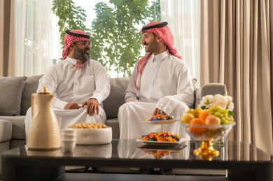 التسامر وتبادل الاحاديث، جلسة عائلية سعيدة، تعابير المودة والمحبة، الاستمتاع بقضاء وقت مع الابناء، رجل كبير بالسن عربي خليجي سعودي يرتدي الثوب التقليدي والشماغ يجلس على الاريكة بجانب ابنه الشاب في غرفة الجلوس