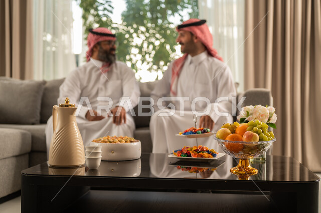 التسامر وتبادل الاحاديث، جلسة عائلية سعيدة، تعابير المودة والمحبة، الاستمتاع بقضاء وقت مع الابناء، رجل كبير بالسن عربي خليجي سعودي يرتدي الثوب التقليدي والشماغ يجلس على الاريكة بجانب ابنه الشاب في غرفة الجلوس