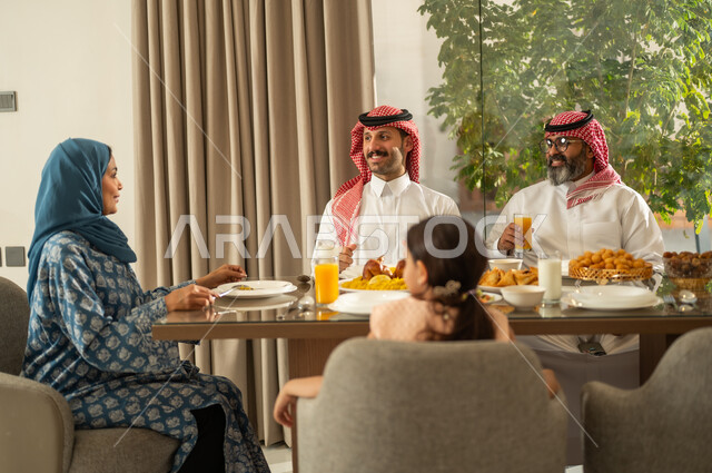 موعد كسر الصيام عند أذان المغرب في رمضان المبارك، فطور رمضاني عائلي، عائلة عربية خليجية سعودية تجلس بايماءات السرور, حسن الضيافة وإكرام الأخرين, أجواء عائلية روحانية دينية إسلامية, اجتماع الأسرة حول مائدة الإفطار المليئة بأصناف المأكولات والمقبلات الشهية