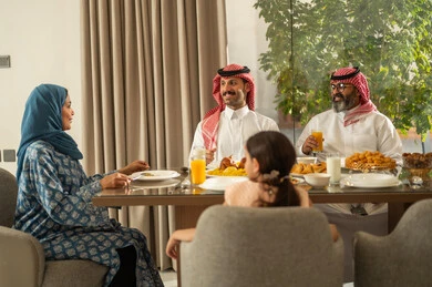 موعد كسر الصيام عند أذان المغرب في رمضان المبارك، فطور رمضاني عائلي، عائلة عربية خليجية سعودية تجلس بايماءات السرور, حسن الضيافة وإكرام الأخرين, أجواء عائلية روحانية دينية إسلامية, اجتماع الأسرة حول مائدة الإفطار المليئة بأصناف المأكولات والمقبلات الشهية
