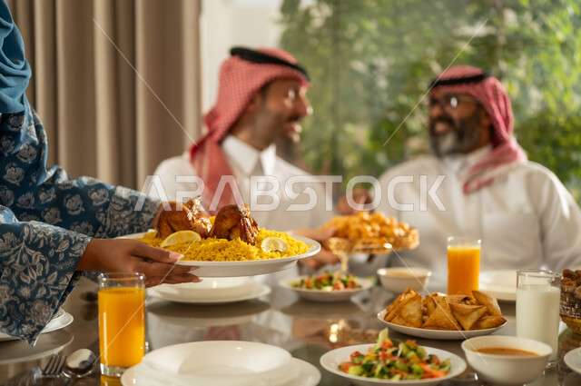 موعد كسر الصيام عند أذان المغرب في رمضان المبارك، فطور رمضاني عائلي، عائلة عربية خليجية سعودية تجلس بايماءات السرور, حسن الضيافة وإكرام الأخرين, أجواء عائلية روحانية دينية إسلامية, اجتماع الأسرة حول مائدة الإفطار المليئة بأصناف المأكولات والمقبلات الشهية
