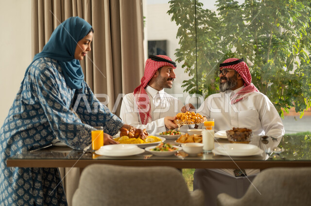 موعد كسر الصيام عند أذان المغرب في رمضان المبارك، فطور رمضاني عائلي، عائلة عربية خليجية سعودية تجلس بايماءات السرور, حسن الضيافة وإكرام الأخرين, أجواء عائلية روحانية دينية إسلامية, اجتماع الأسرة حول مائدة الإفطار المليئة بأصناف المأكولات والمقبلات الشهية