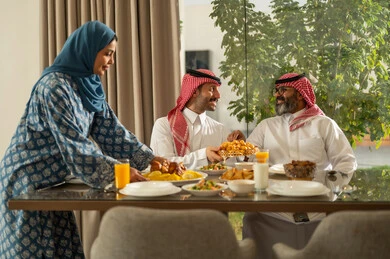 موعد كسر الصيام عند أذان المغرب في رمضان المبارك، فطور رمضاني عائلي، عائلة عربية خليجية سعودية تجلس بايماءات السرور, حسن الضيافة وإكرام الأخرين, أجواء عائلية روحانية دينية إسلامية, اجتماع الأسرة حول مائدة الإفطار المليئة بأصناف المأكولات والمقبلات الشهية