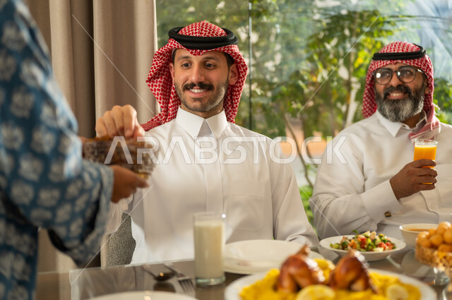Breaking the fast with dates at the Maghrib call to prayer in the holy month of Ramadan, a family Ramadan breakfast, a Saudi Arabian Gulf family sitting with gestures of joy, good hospitality and honoring others, a spiritual, religious, Islamic family atmosphere, the family gathering around the breakfast table filled with delicious food and appetizers