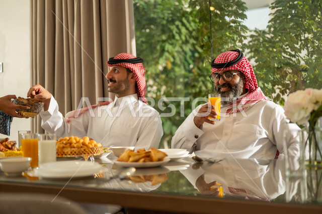  كسر الصيام بالتمر عند أذان المغرب في رمضان المبارك، فطور رمضاني عائلي، عائلة عربية خليجية سعودية تجلس بايماءات السرور, حسن الضيافة وإكرام الأخرين, أجواء عائلية روحانية دينية إسلامية, اجتماع الأسرة حول مائدة الإفطار المليئة بأصناف المأكولات والمقبلات الشهية
