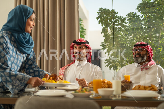 موعد كسر الصيام عند أذان المغرب في رمضان المبارك، فطور رمضاني عائلي، عائلة عربية خليجية سعودية تجلس بايماءات السرور, حسن الضيافة وإكرام الأخرين, أجواء عائلية روحانية دينية إسلامية, اجتماع الأسرة حول مائدة الإفطار المليئة بأصناف المأكولات والمقبلات الشهية