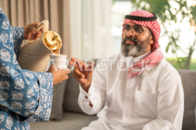 أصالة عادات وتقاليد المجتمع السعودي في المناسبات الاسلامية, كرم الضيافة وحسن الاستقبال في السعودية, إمرأة عربية خليجية سعودية مبتسمة ترتدي العباءة والحجاب تقوم بصب القهوة الخليجية وتقديمها للضيوف في صالة المنزل, تقديم المشروبات الساخنة العربية الأصيلة في الأعياد