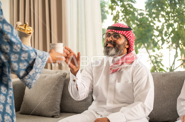 أصالة عادات وتقاليد المجتمع السعودي في المناسبات الاسلامية, كرم الضيافة وحسن الاستقبال في السعودية, إمرأة عربية خليجية سعودية مبتسمة ترتدي العباءة والحجاب تقوم بصب القهوة الخليجية وتقديمها للضيوف في صالة المنزل, تقديم المشروبات الساخنة العربية الأصيلة في الأعياد