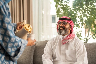 Authentic Saudi society customs and traditions on Islamic occasions, generous hospitality and warm welcome in Saudi Arabia, a smiling Saudi Arabian Gulf woman wearing an abaya and hijab pouring Gulf coffee and serving it to guests in the living room, serving authentic Arabic hot drinks on holidays