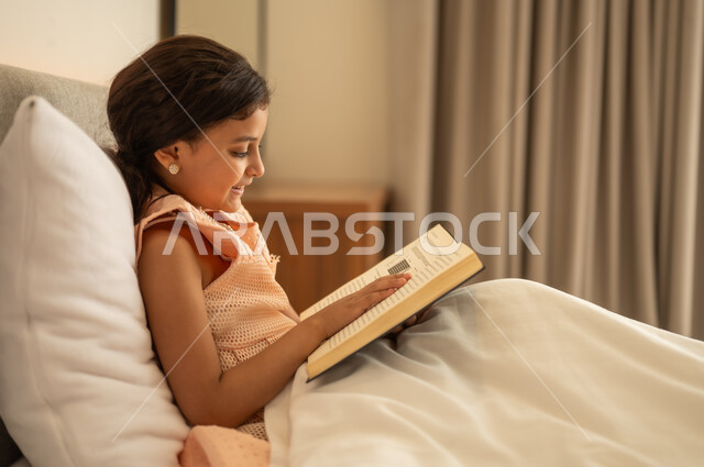Developing children's reading skills, interest in browsing useful books, spending free time learning and self-education, gestures of attention and focus while reading, a close-up photo from the side of a Saudi Arabian Gulf girl sitting on the bed reading carefully, reading entertaining picture stories