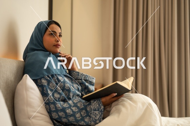 تمضية أوقات الفراغ بالتعلم والتثقف الذاتي, إيماءات الانتباه والتركيز بالمطالعة, امراة عربية خليجية سعودية محجبة ترتدي العباءة تجلس على السرير تقوم بالقراءة بتمعن قبل النوم , مطالعة القصص التصويرية المسلية, تنمية مهارات القراءة, الاهتمام بتصفح الكتب المفيدة
