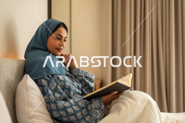 تمضية أوقات الفراغ بالتعلم والتثقف الذاتي, إيماءات الانتباه والتركيز بالمطالعة, امراة عربية خليجية سعودية محجبة ترتدي العباءة تجلس على السرير تقوم بالقراءة بتمعن قبل النوم , مطالعة القصص التصويرية المسلية, تنمية مهارات القراءة, الاهتمام بتصفح الكتب المفيدة