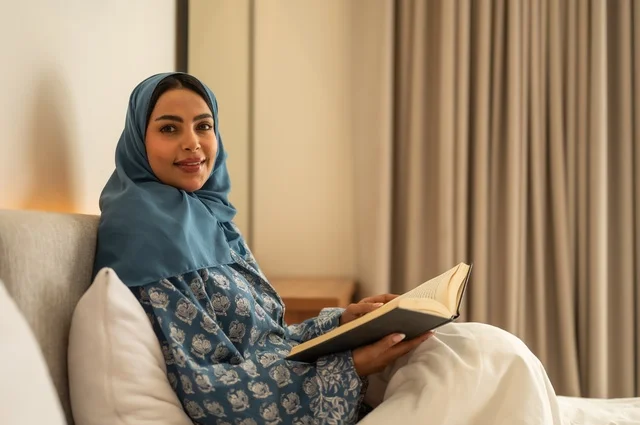 Spending free time learning and self-education, gestures of attention and focus in reading, a veiled Saudi Arabian Gulf woman wearing an abaya sitting on the bed reading carefully before sleeping, reading entertaining picture stories, developing reading skills, interest in browsing useful books