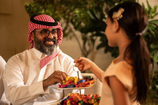 حسن استقبال وإكرام الضيف، صلة الرحم والترابط الاسري, فرحة وبهجة الابناء في ايام العيد السعيد, التجمعات والزيارات الاسرية في الأعياد والمناسبات الاسلامية, رجل عربي خليجي سعودي يرتدي الثوب التقليدي يجلس مع أحفاده بإيماءات الفرح والسرور، ضيافة وحلوى العيد
