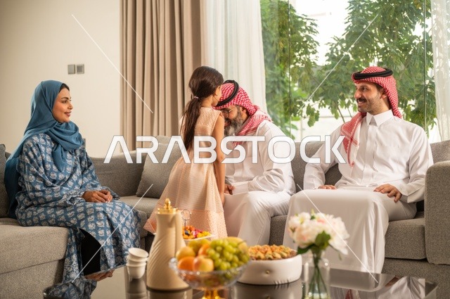 ضيافة وحلوى العيد، حسن استقبال وإكرام الضيف، صلة الرحم والترابط الاسري, فرحة وبهجة الابناء في ايام العيد السعيد, التجمعات والزيارات الاسرية في الأعياد والمناسبات الاسلامية, رجل عربي خليجي سعودي يرتدي الثوب التقليدي يجلس مع أحفاده بإيماءات الفرح والسرور