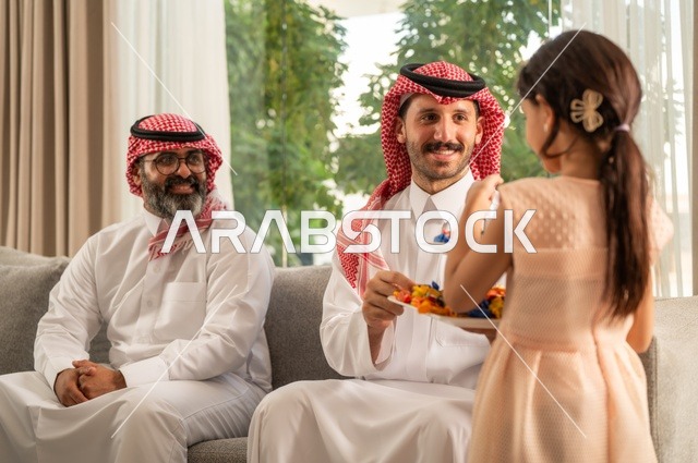 ضيافة وحلوى العيد، حسن استقبال وإكرام الضيف، صلة الرحم والترابط الاسري, فرحة وبهجة الابناء في ايام العيد السعيد, التجمعات والزيارات الاسرية في الأعياد والمناسبات الاسلامية, رجل عربي خليجي سعودي يرتدي الثوب التقليدي يجلس مع أحفاده بإيماءات الفرح والسرور
