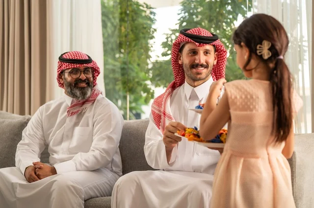 ضيافة وحلوى العيد، حسن استقبال وإكرام الضيف، صلة الرحم والترابط الاسري, فرحة وبهجة الابناء في ايام العيد السعيد, التجمعات والزيارات الاسرية في الأعياد والمناسبات الاسلامية, رجل عربي خليجي سعودي يرتدي الثوب التقليدي يجلس مع أحفاده بإيماءات الفرح والسرور