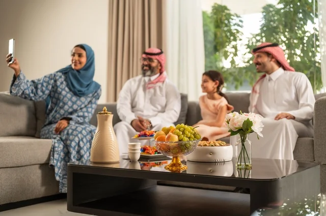 Taking a selfie with a mobile phone, Eid hospitality and sweets, welcoming and honoring guests, joy and happiness of children during the happy Eid days, family gatherings and visits during Islamic holidays and occasions, a Saudi Arabian Gulf man wearing traditional clothing sitting with his grandchildren with gestures of joy and happiness
