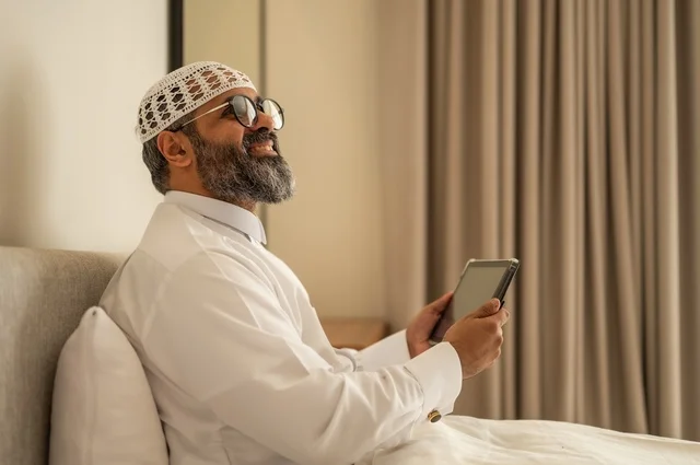 إدارة شؤون الأعمال وتنظيمها عبر الاجهزة التقنية، مهنة ووظيفة ادارية، دمج العمل مع التكنولوجيا، رجل عربي خليجي سعودي يرتدي الثوب التقليدي والقبعة البيضاء يجلس على السرير يعمل على الجهاز اللوحي، استخدام جهاز تقني حديث ومتطور، العمل عن بعد وانجاز المهام المطلوبة