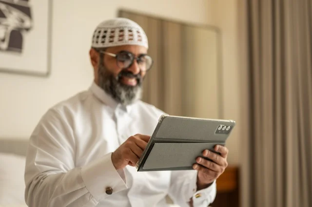 العمل عن بعد وانجاز المهام المطلوبة، إدارة شؤون الأعمال وتنظيمها عبر الاجهزة التقنية، مهنة ووظيفة ادارية، دمج العمل مع التكنولوجيا، رجل عربي خليجي سعودي يرتدي الثوب التقليدي والقبعة البيضاء يجلس على السرير يعمل على الجهاز اللوحي، استخدام جهاز تقني حديث ومتطور