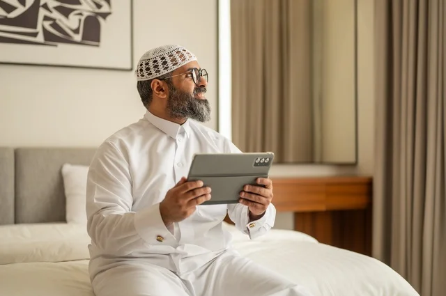 Working remotely and completing required tasks, managing and organizing business affairs through technical devices, an administrative profession and job, integrating work with technology, a Saudi Arabian Gulf man wearing traditional thobe and a white hat sitting on the bed working on a tablet, using a modern and advanced technical device