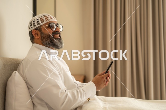 تصفح مواقع التواصل الاجتماعي، رجل عربي خليجي سعودي يرتدي الثوب التقليدي والقبعة البيضاء يجلس على السرير يمسك بيده الهاتف المحمول، الدردشة والتواصل مع الأهل و الأصدقاء، استخدام جهاز تقني حديث ومتطور، العمل عن بعد وانجاز المهام المطلوبة