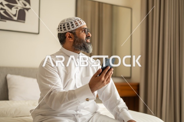 تصفح مواقع التواصل الاجتماعي، رجل عربي خليجي سعودي يرتدي الثوب التقليدي والقبعة البيضاء يجلس على السرير يمسك بيده الهاتف المحمول، الدردشة والتواصل مع الأهل و الأصدقاء، استخدام جهاز تقني حديث ومتطور، العمل عن بعد وانجاز المهام المطلوبة
