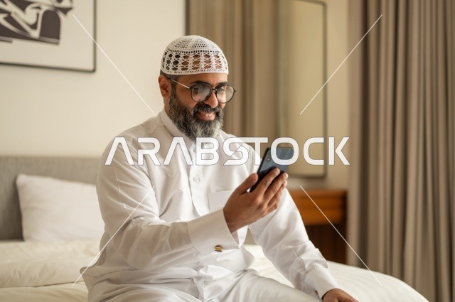 Browsing social media, a Saudi Arabian Gulf man wearing traditional thobe and white hat sitting on the bed holding a mobile phone, chatting and communicating with family and friends, using a modern and advanced technical device, working remotely and completing the required tasks