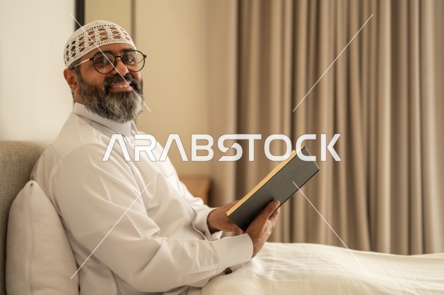 قضاء أوقات الفراغ بالتثقيف والتعلم, تنمية مهارات القراءة, الاهتمام بتصفح الكتب المفيدة، رجل عربي خليجي سعودي يرتدي الثوب التقليدي والقبعة البيضاء يجلس على السرير في المنزل ويحمل بيده كتاب