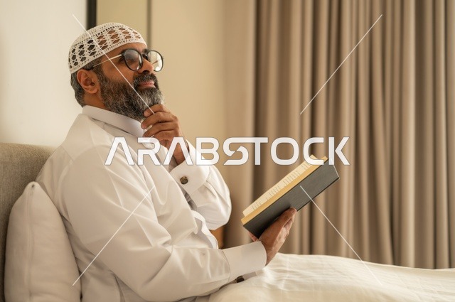قضاء أوقات الفراغ بالتثقيف والتعلم, تنمية مهارات القراءة, الاهتمام بتصفح الكتب المفيدة، رجل عربي خليجي سعودي يرتدي الثوب التقليدي والقبعة البيضاء يجلس على السرير في المنزل ويحمل بيده كتاب