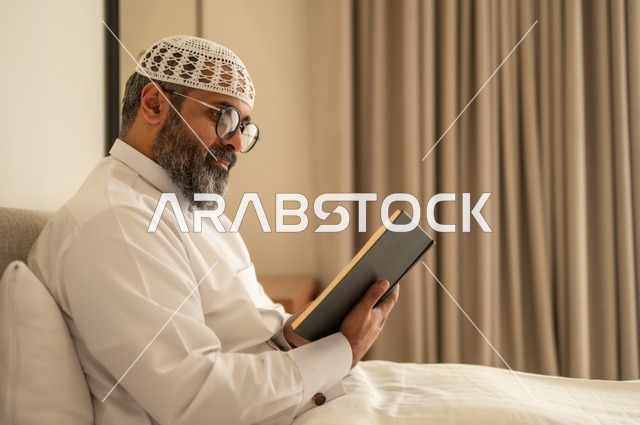 قضاء أوقات الفراغ بالتثقيف والتعلم, تنمية مهارات القراءة, الاهتمام بتصفح الكتب المفيدة، رجل عربي خليجي سعودي يرتدي الثوب التقليدي والقبعة البيضاء يجلس على السرير في المنزل ويحمل بيده كتاب