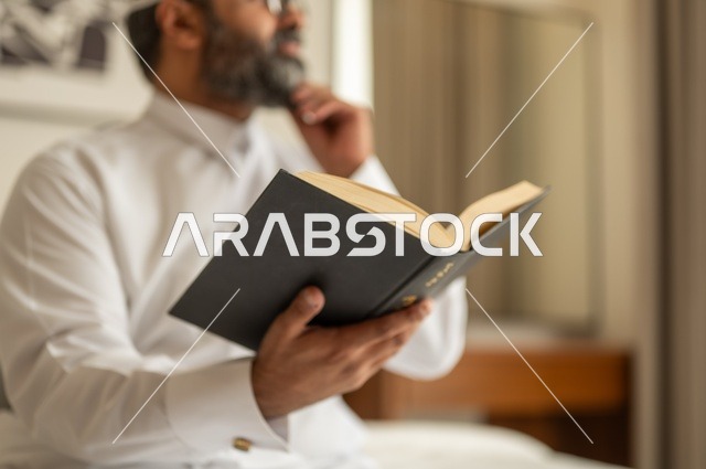 قضاء أوقات الفراغ بالتثقيف والتعلم, تنمية مهارات القراءة, الاهتمام بتصفح الكتب المفيدة، رجل عربي خليجي سعودي يرتدي الثوب التقليدي والقبعة البيضاء يجلس على السرير في المنزل ويحمل بيده كتاب