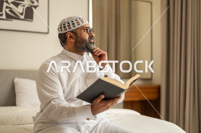 قضاء أوقات الفراغ بالتثقيف والتعلم, تنمية مهارات القراءة, الاهتمام بتصفح الكتب المفيدة، رجل عربي خليجي سعودي يرتدي الثوب التقليدي والقبعة البيضاء يجلس على السرير في المنزل ويحمل بيده كتاب