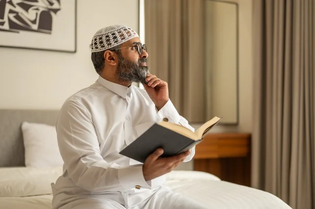 قضاء أوقات الفراغ بالتثقيف والتعلم, تنمية مهارات القراءة, الاهتمام بتصفح الكتب المفيدة، رجل عربي خليجي سعودي يرتدي الثوب التقليدي والقبعة البيضاء يجلس على السرير في المنزل ويحمل بيده كتاب
